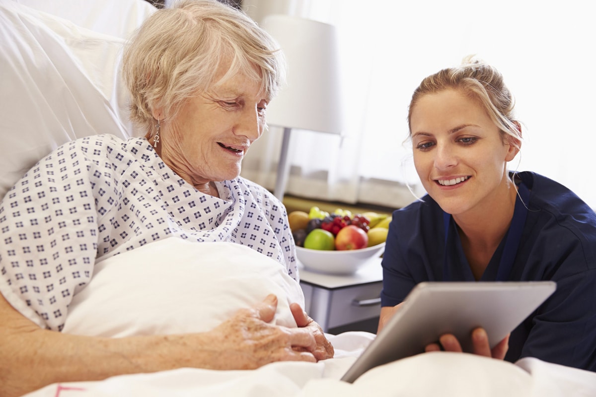 PACS-300x200-computer1 A caregiver wearing blue scrubs showing a computer to an elderly woman in bed
